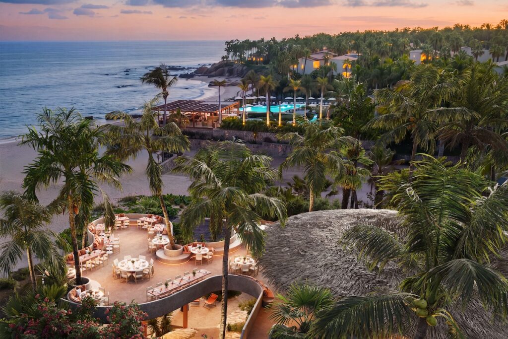 Elevated view of a tropical beachfront resort with an outdoor dining area, palm trees, swimming pool, and ocean at sunset.