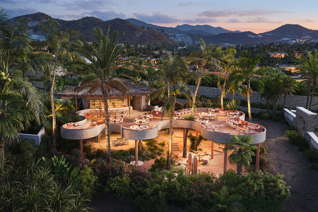 Aerial view of an open-air tropical restaurant surrounded by palm trees and lush landscaping, with mountains in the background at sunset.