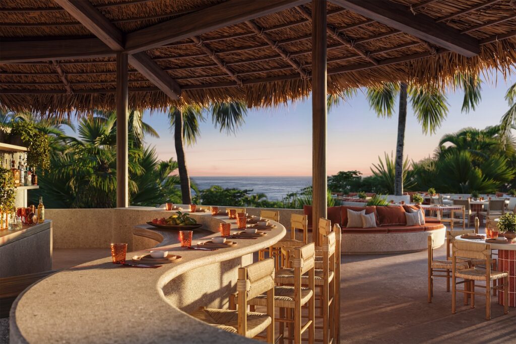 Open-air restaurant and bar beneath a thatched roof, featuring curved counters, natural wood seating, tropical greenery, and ocean views.