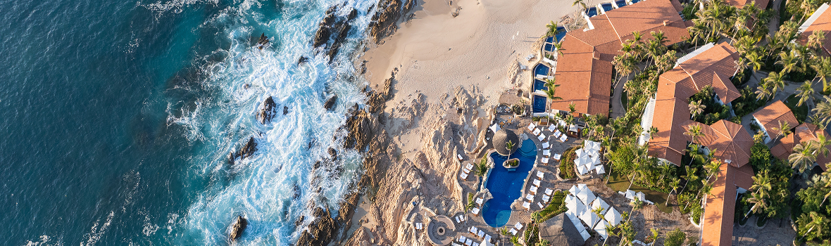 Aerial view of a beachfront resort with terracotta-roofed buildings, palm trees, and pools set along a rocky coastline, where waves crash against the shore beside a sandy beach.