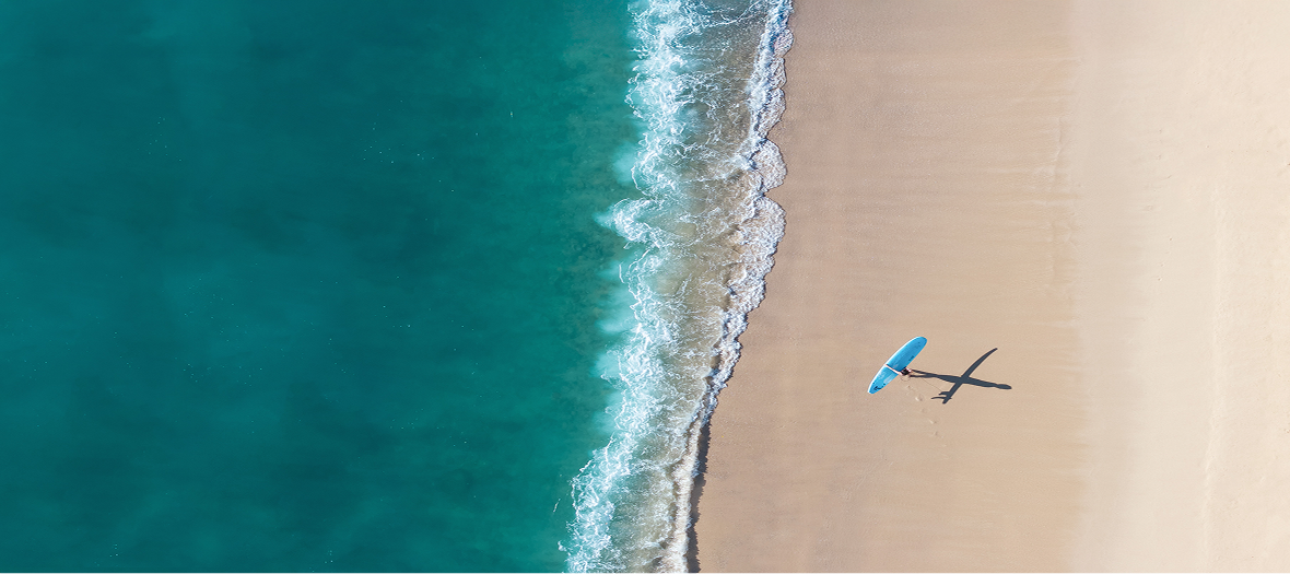 Aerial view of a lone surfer carrying a blue surfboard along a pristine sandy beach, with turquoise waves gently breaking along the shoreline.