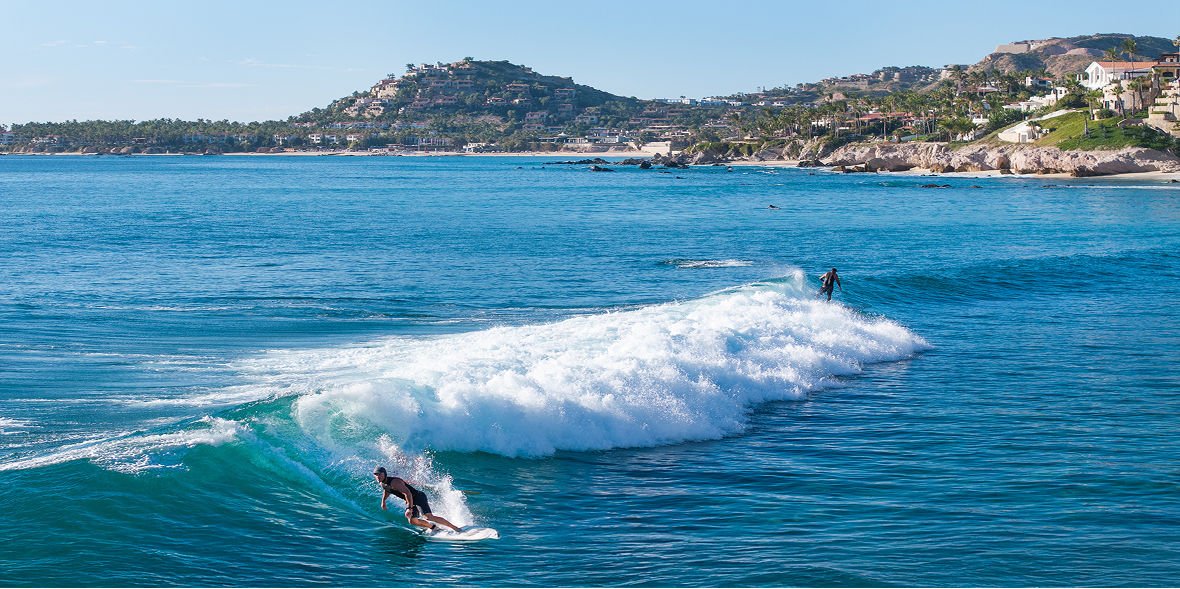 Surfers riding rolling ocean waves along a scenic coastline, with clear blue water in the foreground and hillside homes and palm trees in the distance.