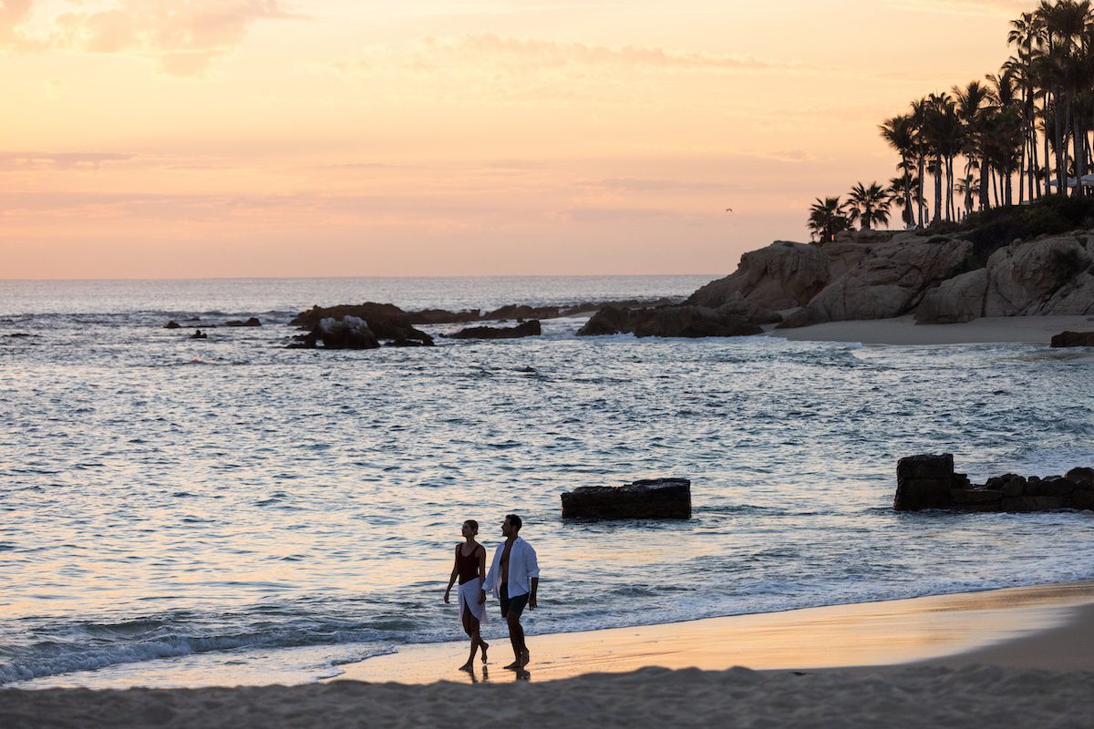 Couple walking along a sandy beach at sunset, with gentle waves, rocky shoreline, and palm trees silhouetted against a warm, glowing sky.