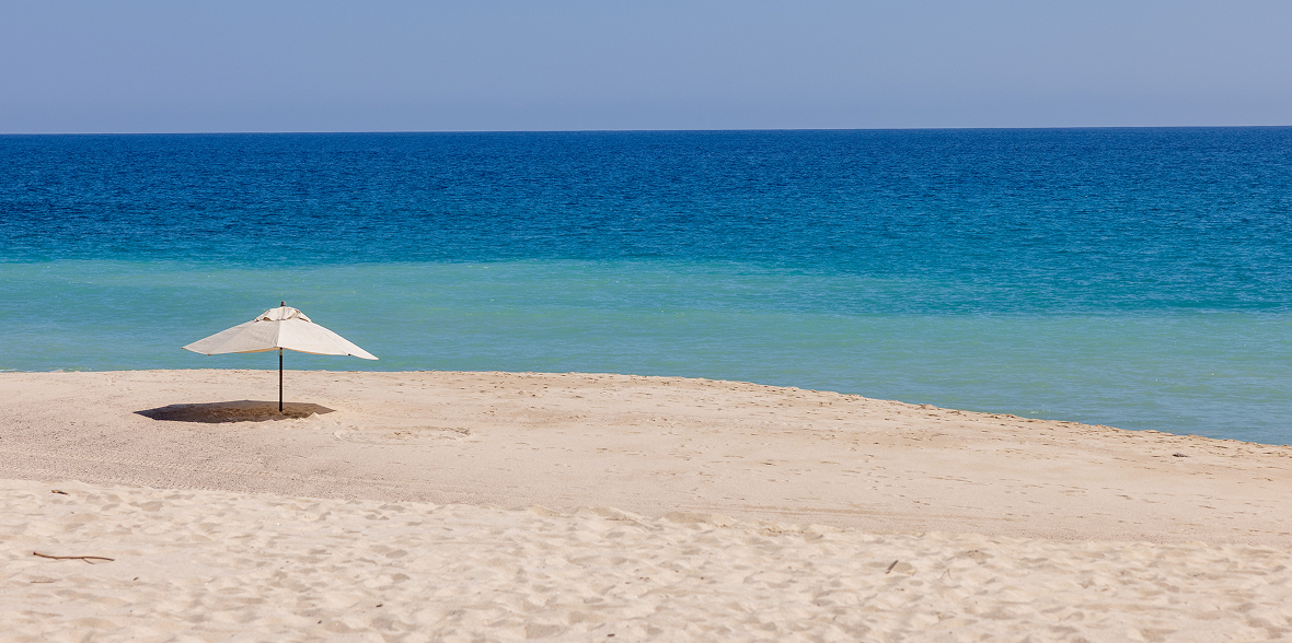 Serene sandy beach with a single white umbrella set near the shoreline, overlooking calm, turquoise ocean waters under a clear sky.