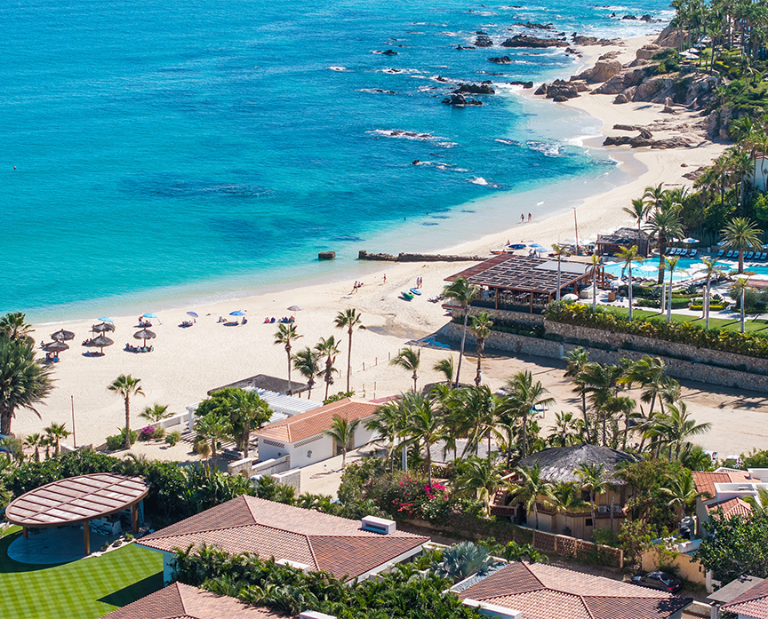 Aerial view of Playa Palmilla featuring turquoise ocean waters, a sandy beach lined with palm trees, and a beachfront resort community with pools, terraces, and coastal homes.