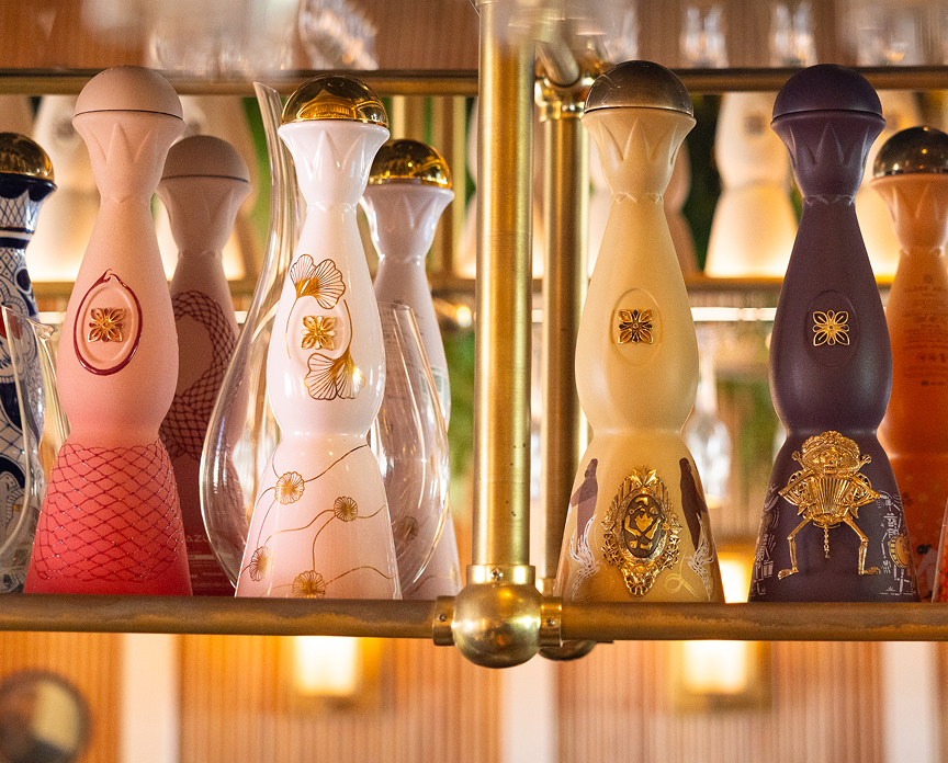 Display of Clase Azul tequila bottles in ornate ceramic decanters with hand-painted designs and gold accents, arranged on a bar shelf in a warm, ambient setting.