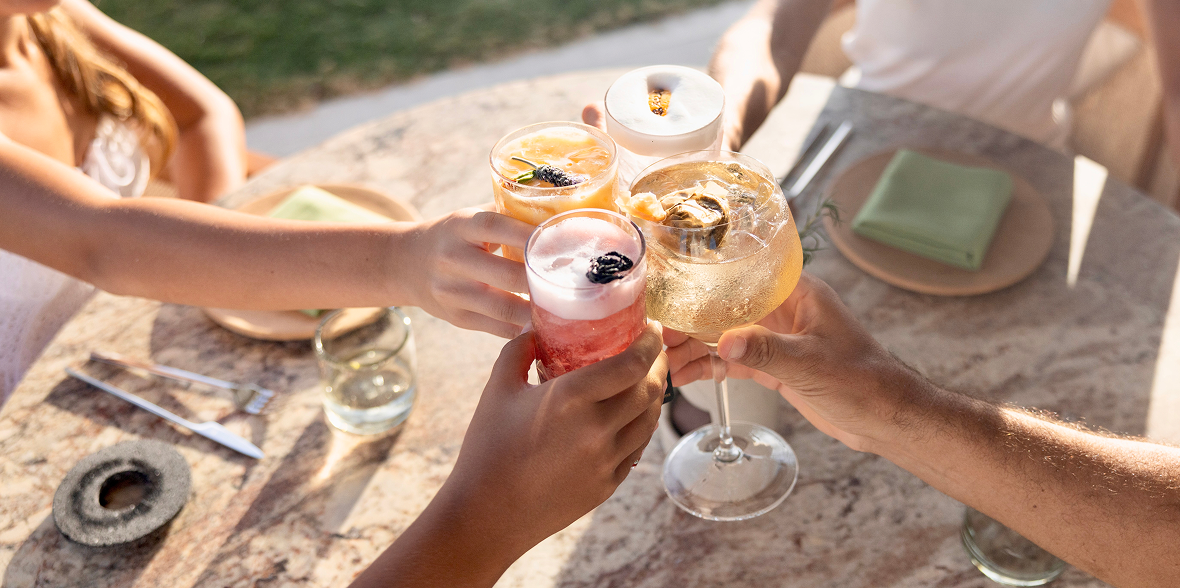 Friends toasting with colorful cocktails over a sunlit outdoor table, capturing a relaxed, social moment with drinks garnished and glowing in warm light.