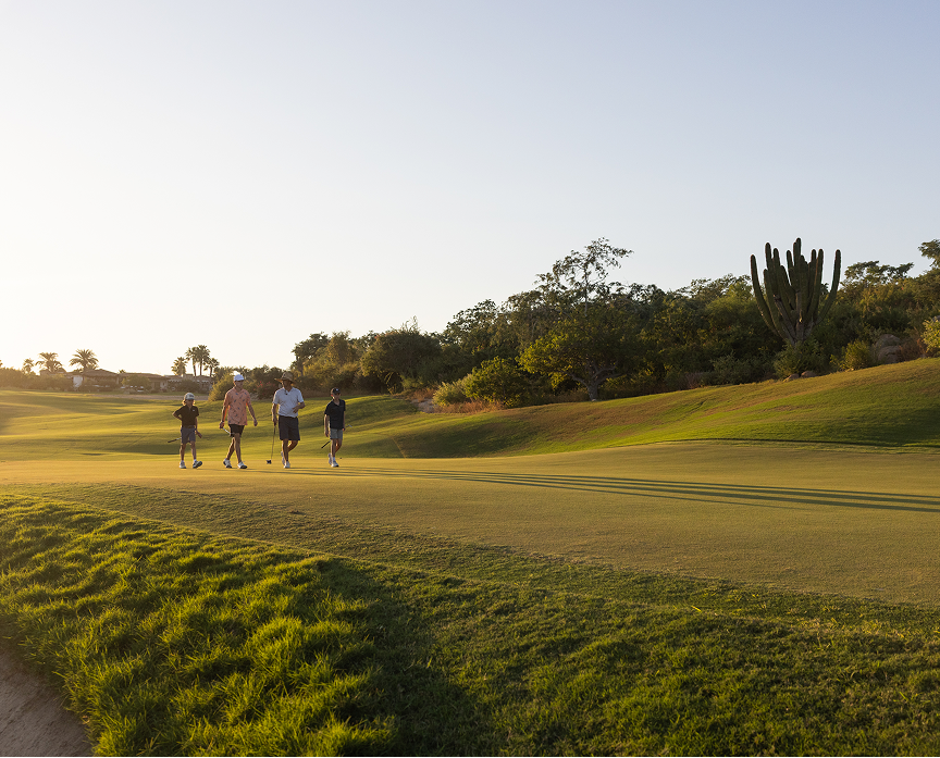Group of golfers walking across a sunlit fairway at golden hour, with long shadows stretching across the green and desert vegetation lining the course.