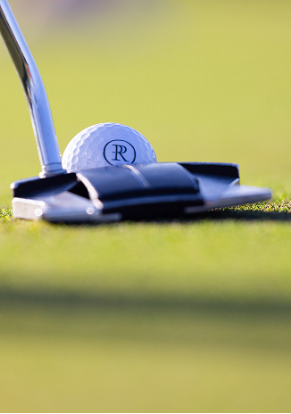 Close-up of a golf ball on green grass beside a putter club, ready for a putt.