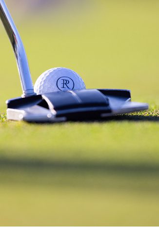 Close-up of a golf putter striking a golf ball marked with a the Palmilla Reserve logo on a smooth green.