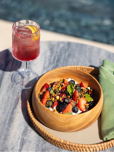 Fresh fruit bowl with berries, coconut, and granola served on a wooden plate beside a red cocktail, set on a marble table near a pool in bright sunlight.