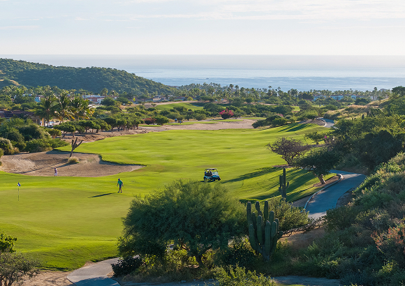 Scenic view of a lush golf course with rolling fairways, sand bunkers, and desert vegetation, with golfers and a cart on the course and the ocean visible in the distance beyond palm-lined hills.