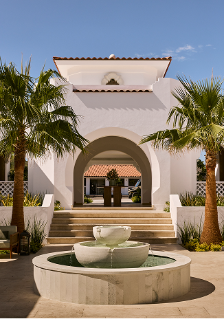White stucco building with arched entryway, palm trees, and a circular fountain in front.