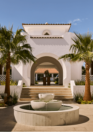 Elegant white stucco entrance with arched architecture, framed by palm trees and centered around a tiered stone fountain, leading into a tranquil courtyard beyond.