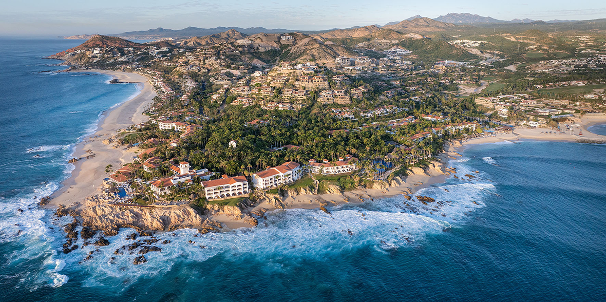 Aerial view of Palmilla set on a rocky point, with terracotta-roofed buildings, palm trees, and sandy beaches surrounded by turquoise ocean, backed by rolling desert mountains.