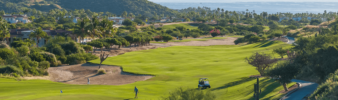 Wide view of a manicured golf course with lush green fairways, sand bunkers, and scattered desert vegetation, with golfers and a cart on the course and palm trees, hillside homes, and the ocean visible in the distance.